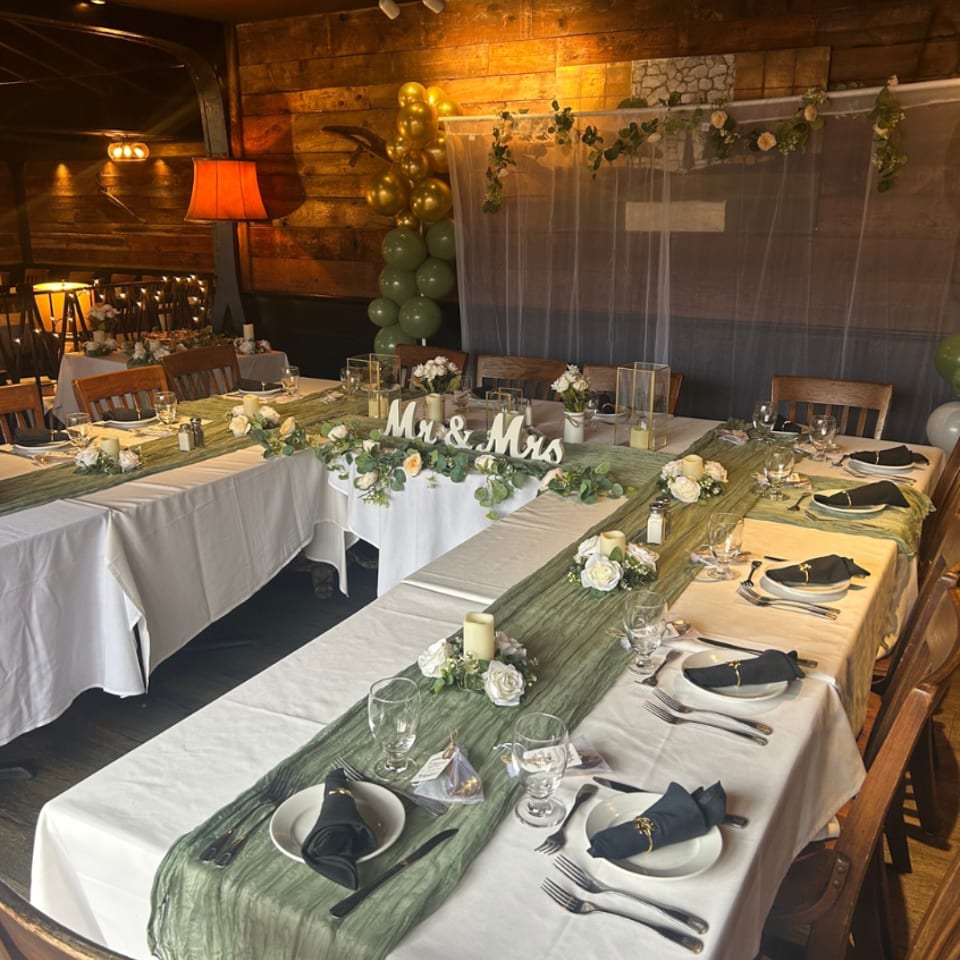Rustic restaurant dining room decorated for a wedding with white tablecloths, green runners, floral centerpieces, and gold-green balloons.
