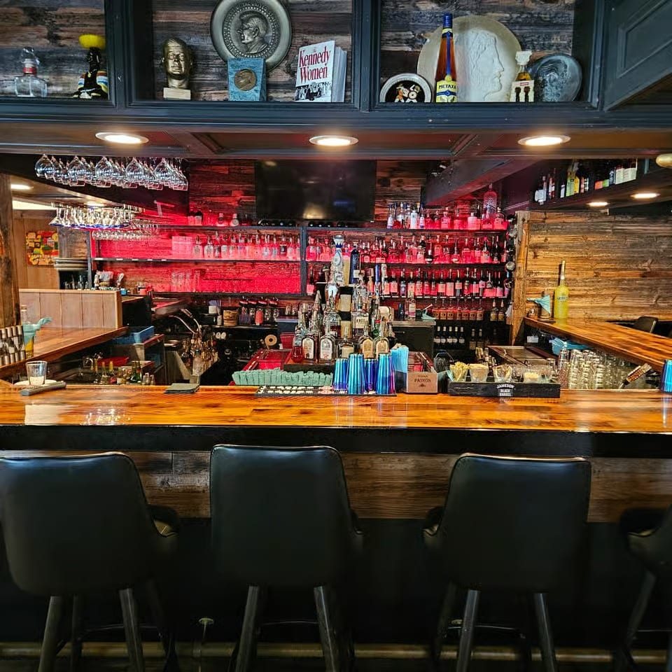 Rustic restaurant bar with polished wooden counter, black chairs, liquor shelves, hanging glassware, and vintage wall decor.